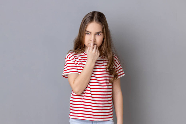 Don't lie. Little girl wearing striped T-shirt looking with incredulous suspicious gaze and touching nose, gesturing you are liar, suspecting falsehood. Indoor studio shot isolated on gray background. - Фото, зображення