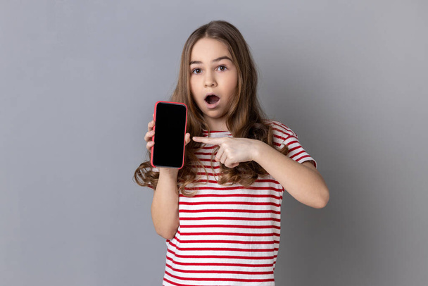 Retrato de una niña sorprendida con una camiseta a rayas apuntando con el dedo al teléfono inteligente con una pantalla blanca vacía, conmocionada por la aplicación. Estudio interior plano aislado sobre fondo gris. - Foto, Imagen