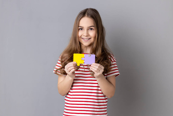 Little girl wearing striped T-shirt holding and showing two jigsaw puzzle pieces, matching and connecting puzzles, solving problems with good result. Indoor studio shot isolated on gray background. - Foto, Imagen