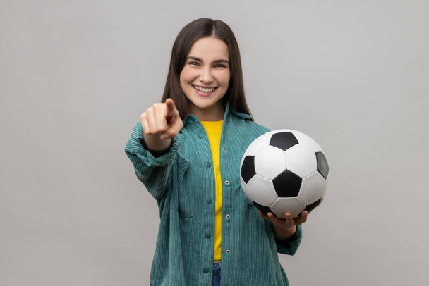 Young beautiful dark haired woman holding soccer ball, looking at camera with toothy smile, pointing finger to camera, wearing casual style jacket. Indoor studio shot isolated on gray background. - Foto, Bild