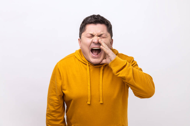 Angry nervous middle aged man loudly yelling widely opening mouth holding hands on face, screaming announcing his opinion, wearing urban style hoodie. Indoor studio shot isolated on white background. - Photo, image
