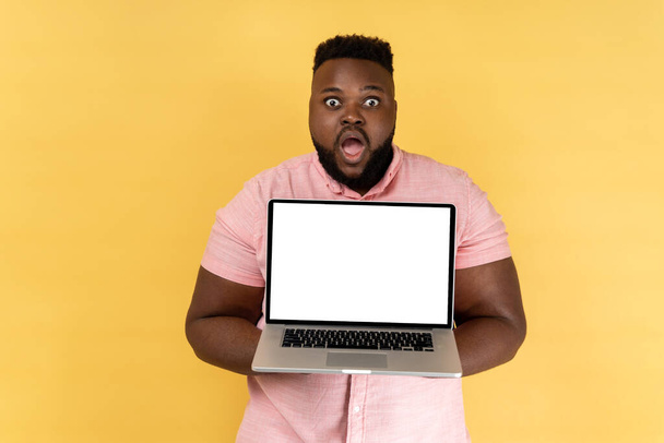 Portrait of shocked amazed man wearing pink shirt holding notebook, showing white blank screen on his laptop, looking at camera with big eyes. Indoor studio shot isolated on yellow background. - Foto, Imagem