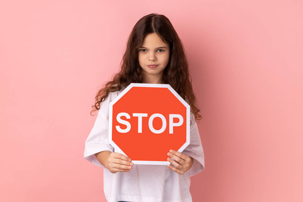 Portrait of strict bossy adorable little girl wearing white T-shirt holding red stop sign looking at camera with serious expression. Indoor studio shot isolated on pink background. - Фото, зображення