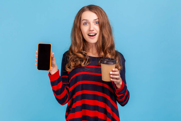 Astonished woman wearing striped casual style sweater, holding coffee to go and showing smart phone with blank screen for advertisement. Indoor studio shot isolated on blue background. - Photo, Image