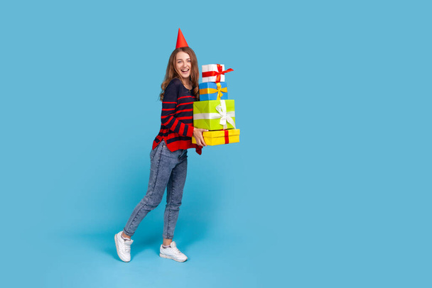 Full length portrait of woman wearing striped casual style sweater and red party box standing with many present boxes, looking at camera with excitement. Indoor studio shot isolated on blue background - Fotó, kép