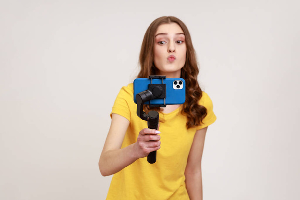 Young attractive woman with camera phone on stabilizer shoots video for her vlog, making kissing gesture, wearing yellow casual style T-shirt. Indoor studio shot isolated on gray background. - Φωτογραφία, εικόνα