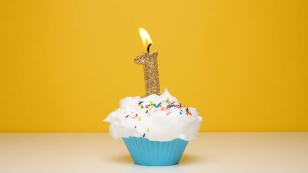 Birthday cupcake With Burning golden Candle number one on bright yellow Background. Slow motion first anniversary video banner with white confetti. - Imágenes, Vídeo