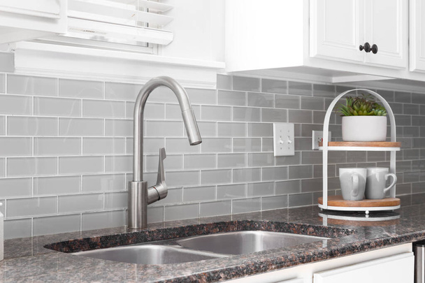 A kitchen sink detail shot with white cabinets, grey subway tile backsplash, and a granite countertop. - Фото, зображення