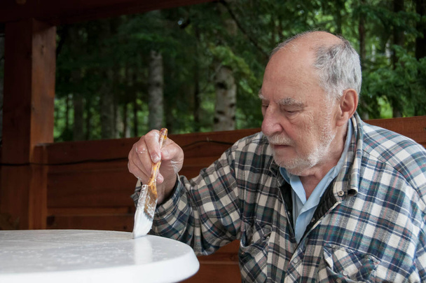 Дедушка с выразительным лицом аккуратно рисует садовый стол - Фото, изображение