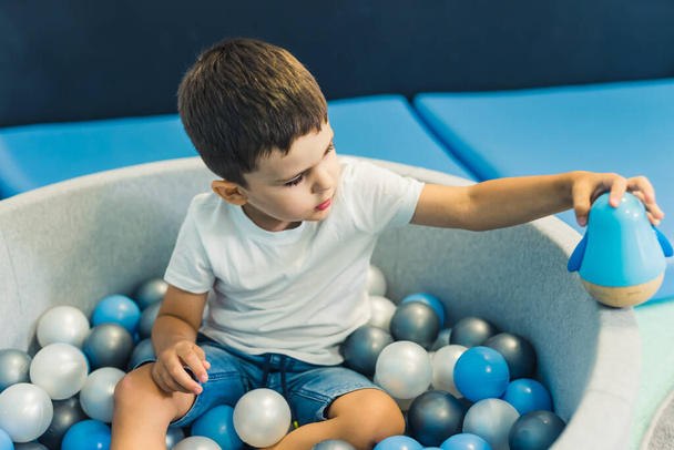 Παιδί παιδί παίζει με ένα παιχνίδι, ενώ κάθεται σε ένα λάκκο μπάλα γεμάτη πολύχρωμες μπάλες. Μια μπάλα pit - ένα εξαιρετικό μέρος για τα παιδιά να πηδούν, συντριβή, και ταλαντεύεται. Αισθητηριακό παιχνίδι στο νηπιαγωγείο. Υψηλή ποιότητα - Φωτογραφία, εικόνα