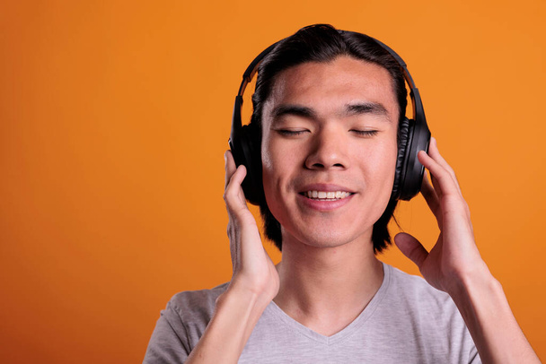 Young adult asian man enjoying music in wireless headphones with satisfied facial expression, close view. Smiling relaxed teenager wearing earphones, listening to song closeup - Foto, imagen