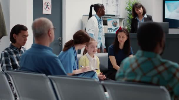 Health specialist talking to small child and mother in hospital reception lobby, taking notes about consultation. Nurse consulting little girl sitting in facility waiting room, healthcare support. - Filmmaterial, Video