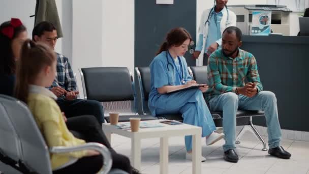 Afrikanisch-amerikanischer Patient berät sich mit Krankenschwester und macht sich Notizen in Wartezimmer-Lobby. Diverse Menschen unterhalten sich über den Besuch von Vorsorgeuntersuchungen und schreiben Berichte. - Filmmaterial, Video
