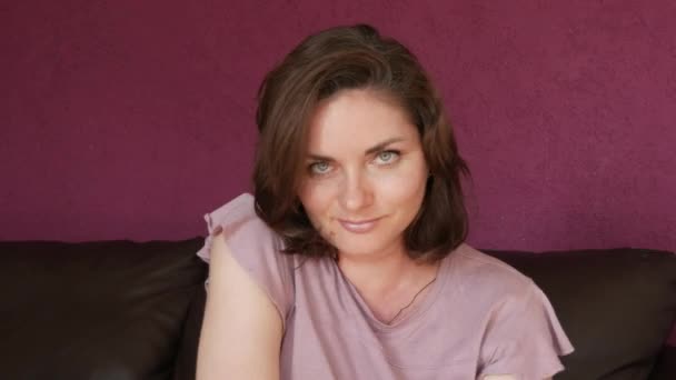 Close-up portrait of a young beautiful woman with green eyes and dark hair, who looks directly into the camera fooling around, grimacing having fun and dancing on a leather sofa in the room. - Video