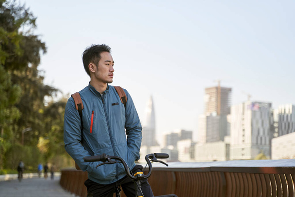 asian young adult man sitting on his bicycle lost in thought in city park - Foto, Imagen