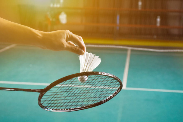 El jugador de bádminton sostiene la raqueta y el volante de crema blanca frente a la red antes de servirlo en otro lado de la cancha. - Foto, Imagen