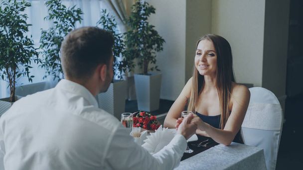 Young attractive girl in fancy clothes is talking to her bearded boyfriend while dining in restaurant. Flowers, champagne glasses, green plants and tableware are visible. - Photo, image