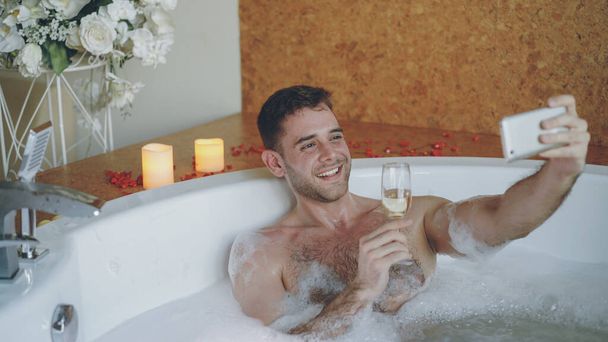 Handsome young man is taking selfie with champagne glass using smartphone in hot tub with foam in modern spa salon. He is smiling and posing looking at camera. - 写真・画像