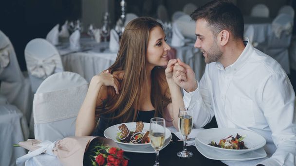 Happy loving couple is holding hands, talking and kissing during romantic dinner in luxurious restaurant. Affectionate relationship, love and fine dining concept. - Фото, зображення