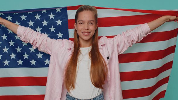 Lovely teenager girl waving and wrapping in American USA flag, celebrating, human rights and freedoms. Independence day. Young stylish child kid isolated alone on blue studio background indoors - Fotoğraf, Görsel