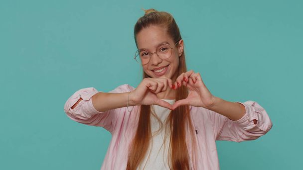 I love you. Smiling lovely pretty teenager girl makes heart gesture demonstrates love sign expresses good feelings and sympathy. Young teen female child kid isolated on blue studio background indoors - Fotografie, Obrázek