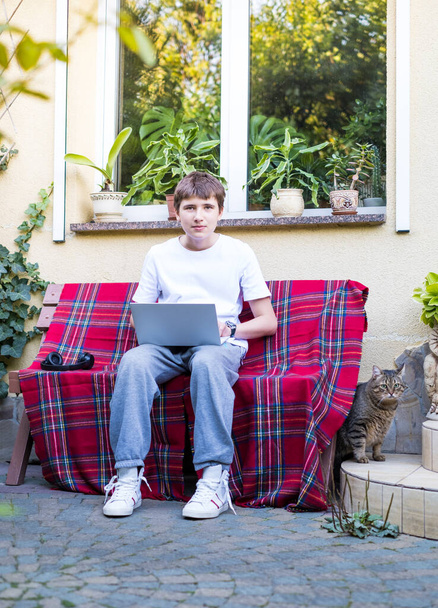 Teenager with laptop on the bench near the house. Technology studying, distance education, e-learning course, virtual lesson, communicates on social networks, studies school materials - Foto, Bild