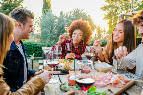 Group of multiethnic friends living healthy lifestyle and smiling and joking while doing aperitif and drinking red wine at outdoor pub restaurant - Foto, Imagem