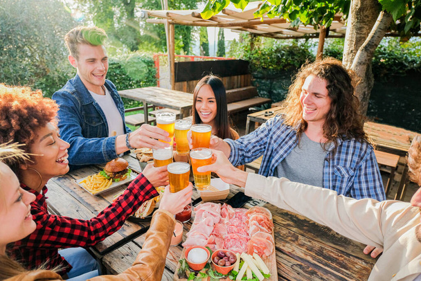 Group of multiethnic friends living healthy lifestyle and smiling and joking while drinking beer at outdoor pub restaurant - Young people toasting with beer glass during happy hour at bar - 写真・画像