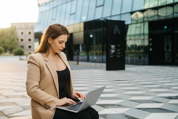 Happy young woman blonde office worker typing on laptop while sitting outdoors in the city, business woman or female freelancer answering emails, working outdoors, cityscape in the background.  - Foto, imagen
