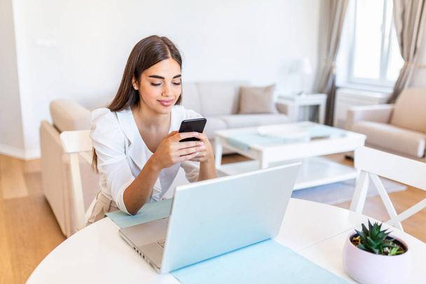 Улыбающаяся молодая женщина учится дома с помощью ноутбука и мобильного телефона, концепция электронного обучения онлайн, молодая деловая женщина, проводящая конференцию с клиентом на ноутбуке. - Фото, изображение