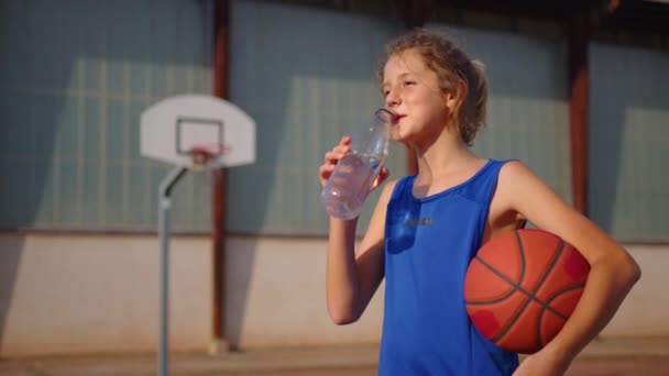 Boy taking break after basketball game and drink fresh water from bottle. Sport concept. Rehydration after basketball game. Basketball player drinking water. Boy in a blue basketball uniform. - Filmmaterial, Video