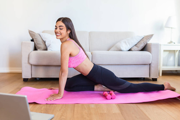 Girl training at home, doing workout and watching videos on laptop, training in living room. Beautiful woman is doing side plank exercise. She is watching video on the internet and repeating the tasks - Photo, Image
