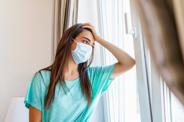 Home quarantine. Caucasian woman sitting at window in a medical mask, looking out, wants to go out. protection against coronavirus infection, pandemics, disease outbreaks and epidemics. - Foto, imagen
