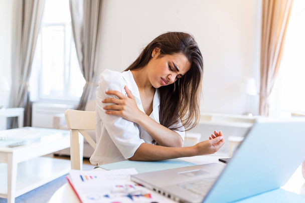 Молодая женщина с руками, держащая боль в плече. Синдром офиса и концепция здравоохранения. Бизнес-женщина, работающая со своим ноутбуком и с болью в плече в домашнем офисе - Фото, изображение