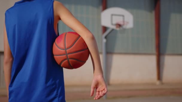 Boy standing on a basketball court and holding a basketball under his arm. Video from the back shot of teenager standing with a ball in his hand and a basketball basket in front of him. - Filmmaterial, Video