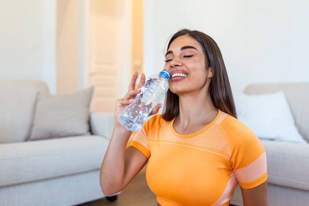 Jovem mulher adulta bebendo água de garrafa de plástico, sentada no tapete de fitness e descansando após o treinamento em casa
 - Foto, Imagem