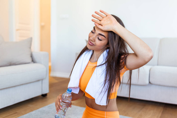 Young adult woman drinking water from plastic bottle, sitting on fitness mat and resting after training at home. Beautiful young woman wiping sweat with a towel, exhausted after the daily training. - Photo, image