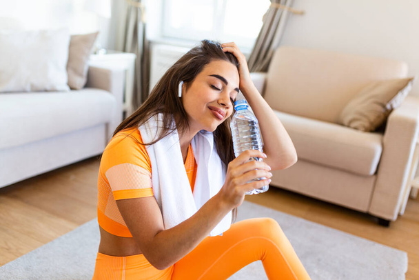 Young adult woman drinking water from plastic bottle, sitting on fitness mat and resting after training at home. Beautiful young woman wiping sweat with a towel, exhausted after the daily training. - Fotó, kép