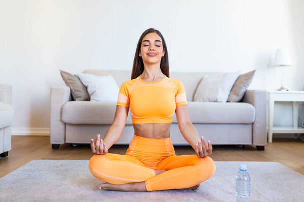 Young attractive smiling woman practicing yoga, sitting in Half Lotus exercise, Ardha Padmasana pose, working out, wearing sportswear, meditation session, indoor full length, home interior - 写真・画像
