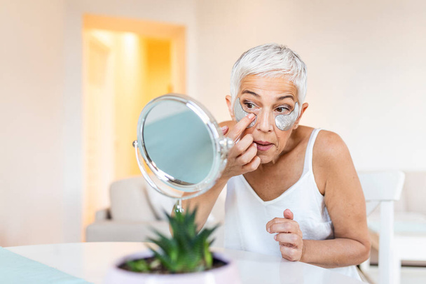 Close up photo of lovely retiree applying nourishment eye mask under eyes while looking in the mirror. Beauty concept - Valokuva, kuva