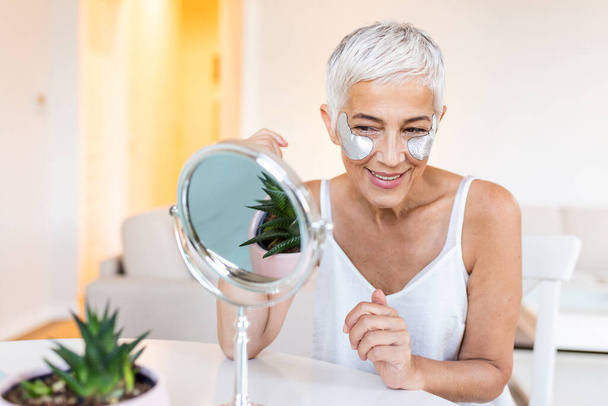 Smiling mature woman applying rejuvenating eye mask on her face skin. Elderly female enjoy making face massage, peeling, put on mask under eyes in front of the mirror - Photo, image