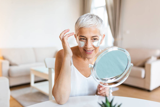Smiling mature woman applying rejuvenating eye mask on her face skin. Elderly female enjoy making face massage, peeling, put on mask under eyes in front of the mirror - 写真・画像
