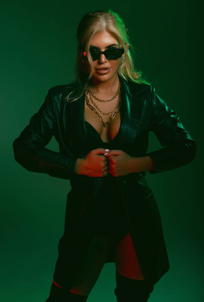 Studio fashion shot of the beautiful rock woman dressed in black leather jacket and black body lingerie, sunglasses on the face. Seductive lady in green light on a dark green background - Zdjęcie, obraz