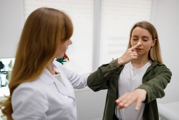 Γυναίκα με κλειστά μάτια αγγίζει τη μύτη με το δάχτυλο κατά τη διάρκεια της φυσικής εξέτασης. Νευρολόγος ελέγχει το συντονισμό των κινήσεων του γυναικείου ασθενούς - Φωτογραφία, εικόνα