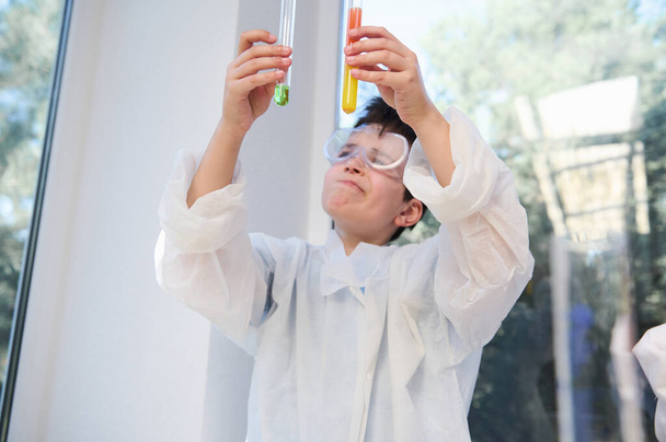 Focus on test tubes with colorful reagents and chemicals in the hands of schoolboy, wearing white lab coat and goggles, doing experiments in chemistry lesson. Back to school. Miracles of chemistry - Photo, Image