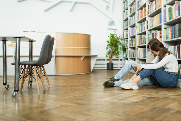 teenage friends sitting on floor near bookshelves and reading book in library - Foto, Imagem