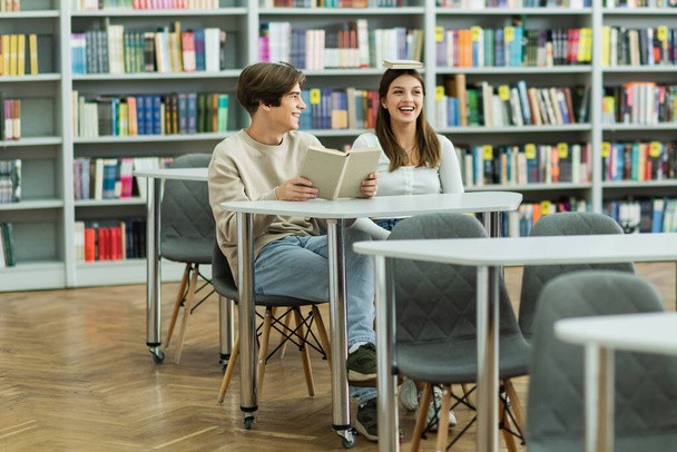 teenage girl with book on head near smiling friend in library reading room - Foto, Bild