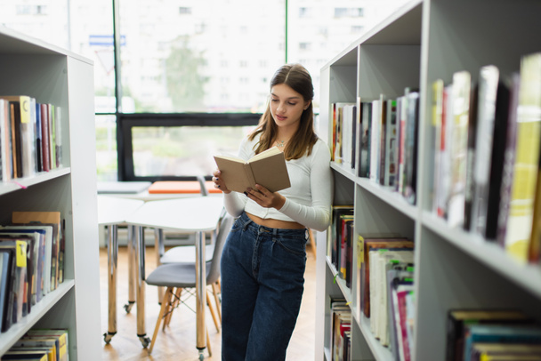 teenage girl standing near shelves in library and reading book - Photo, Image