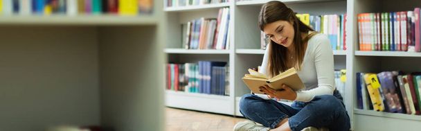 teen student sitting on floor in library and reading book on blurred foreground, banner - Foto, Imagen