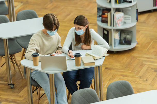 высокий угол обзора подростков в медицинских масках, пишущих возле ноутбука и бумажных стаканчиков в библиотеке - Фото, изображение